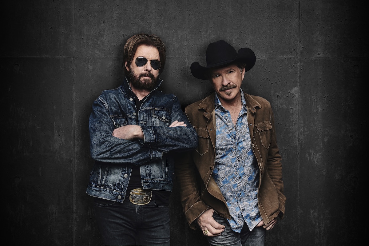 Country music duo Brooks & Dunn pose against a dark wall; Kix Brooks wears a cowboy hat and Ronnie Dunn wears sunglasses in a denim jacket.